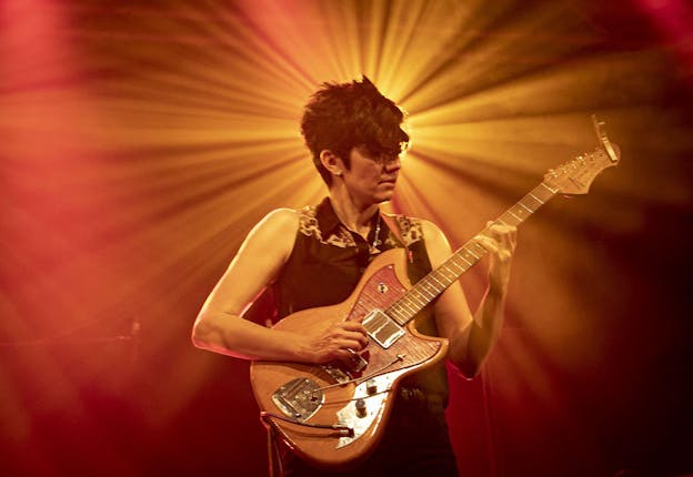 Ava Mendoza plays an electric guitar on a brightly lit stage. They are visible from the hips up, centered with their head in front of an orange, overhead stage light, its bright rays fan out around their head, filling the majority of the image background. Their body is facing the camera while their face is turned to look at their left hand as they press the guitar strings, their right hand rests over the strings. Mendoza is wearing black jeans, a black sleeveless button up blouse with leopard print highlights on the shoulders, a chain necklace, and small earrings.