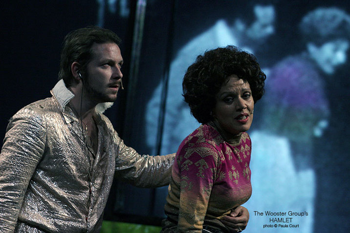 Kate Valk performs, looking distressed and holding her stomach as a man wearing a silver shirt appears behind her. Behind both of them, a blue hued projection of people is on the wall. 