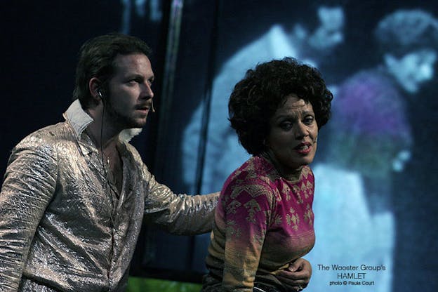 Kate Valk performs, looking distressed and holding her stomach as a man wearing a silver shirt appears behind her. Behind both of them, a blue hued projection of people is on the wall. 