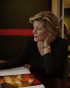 Profile portrait of Agnes Gund with blonde short hair, dressed in a black shirt and gold watch on the left arm. Agnes Gund sits on a table looking into an open book with the picture of a figure with their face cut by the page, dressed in a red and gold gown with jewelry – the other page consists of text on a white background. 