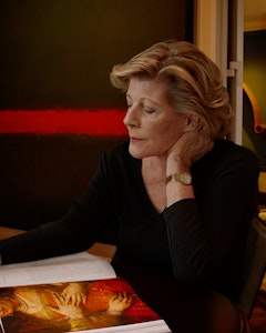 Profile portrait of Agnes Gund with blonde short hair, dressed in a black shirt and gold watch on the left arm. Agnes Gund sits on a table looking into an open book with the picture of a figure with their face cut by the page, dressed in a red and gold gown with jewelry – the other page consists of text on a white background. 