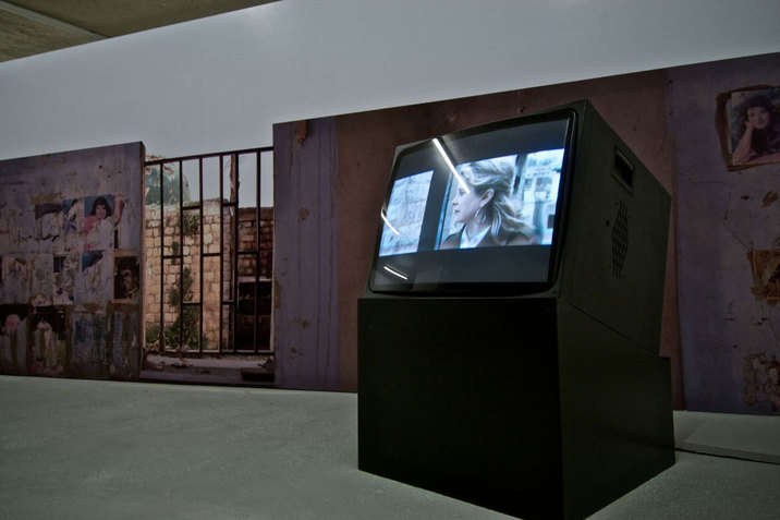 An asymmetrical black slanted box hosts a television displaying a bluish-hued person's face, propped in front of an earthy purple and red wall with a gate revealing a wall of stacked bricks in the distance.