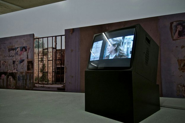 An asymmetrical black slanted box hosts a television displaying a bluish-hued person's face, propped in front of an earthy purple and red wall with a gate revealing a wall of stacked bricks in the distance.