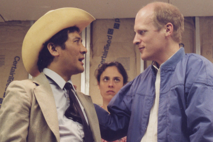 A close up image of three performers in front of a wall made of patterned brown paper. Two performers in the foreground appear deep in conversation. The performer on the left wears a grey suit and a cowboy hat. The performer on the right wears a blue jacket and a white shirt. Behind them, a performer looks on with concern. 