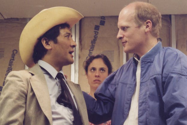 A close up image of three performers in front of a wall made of patterned brown paper. Two performers in the foreground appear deep in conversation. The performer on the left wears a grey suit and a cowboy hat. The performer on the right wears a blue jacket and a white shirt. Behind them, a performer looks on with concern. 