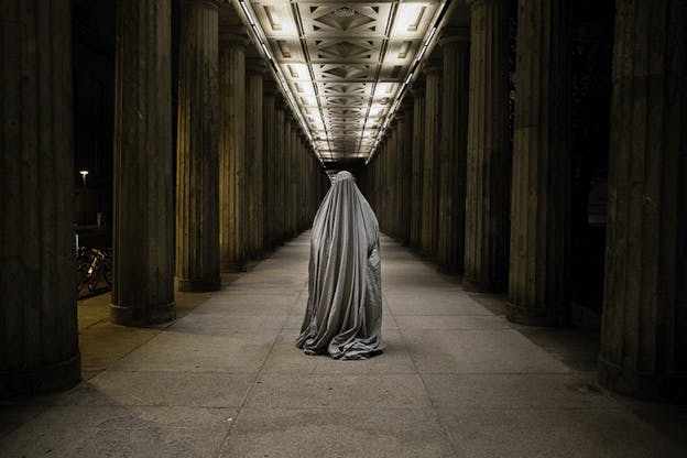 A form covered by a silver sheet stands in a hallway lined with columns. The ceiling consists of lighted geometrical shapes.