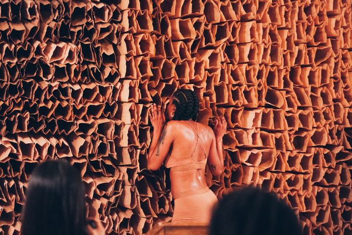 A performer dressed in nude undewear has placed their face inside one of the multiple brown paperbags that have been put together forming a wall.