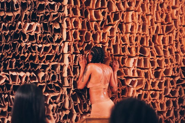 A performer dressed in nude undewear has placed their face inside one of the multiple brown paperbags that have been put together forming a wall.