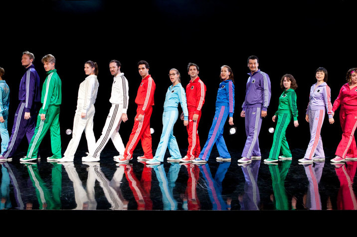 On a dark stage, a spotlit line of performers dressed in varying monochromatic Adidas tracksuits and matching Converse stand sideways and face forwards, one leg outstretched in front of the other.