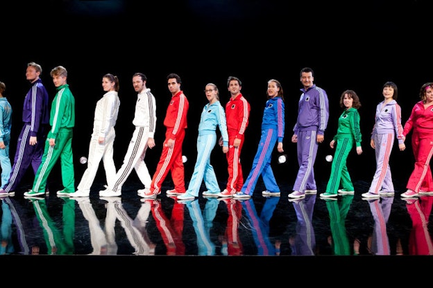 On a dark stage, a spotlit line of performers dressed in varying monochromatic Adidas tracksuits and matching Converse stand sideways and face forwards, one leg outstretched in front of the other.