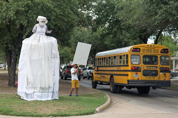 A white dressed tall figure held upwards by a structure under their skirts stands next to a person holding a sign and looking towards a school bus.