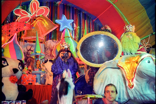 There is a densely crowded space filled with plush toys, sparkly decorations, and neon lights. The ceiling of this space is curved and colored with thick colorful stripes. The back wall of this space is covered in multi colored ribbons. In the center of the space, a person wearing a purple outfit with sparkles on it and a crown holds a camera to their face, obscuring it. Behind them, another person holds a mirror with a gold frame that seems to reflect a person taking a flash photograph. 
