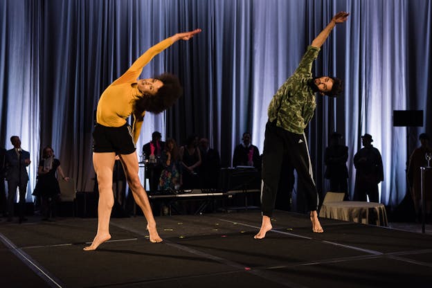 Two performers on a black stage mid-air with each their right hand looping over their head.