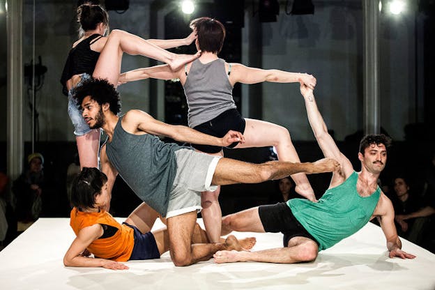Performers in tank tops and shorts twist their bodies together by striking varied poses with a foot and hand touching another. Behind the elevated and illuminated stage, a dim audience observes.