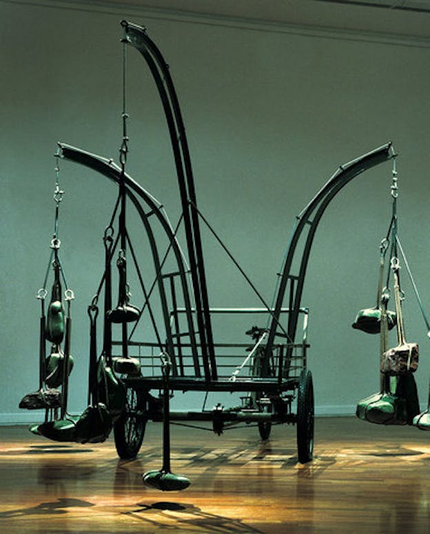 A 45-degree view of a mixed media installation made of found and recycled objects. A tricycle is used as the base to support one longer and two shorter curved iron structures branching outward, each with several river stones are hung at the end. The installation sits on a wooden floor against a palladian blue background with natural lighting coming from above.