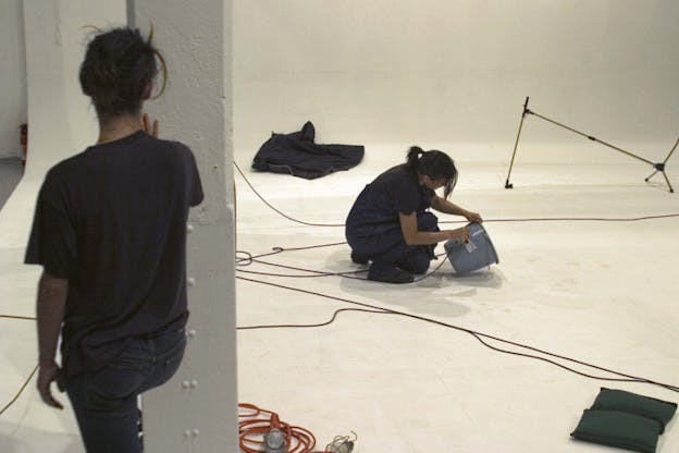 A performance still of two performers in a white studio. On the floor, there are black wires, a red wire attached to a lightbulb, and a navy blanket. The performers are dressed in navy t-shirts and blue jeans with their hair in pony tails. On performer crouches on the floor, writing something on a grey bucket while the other performer watches from behind a white column. 