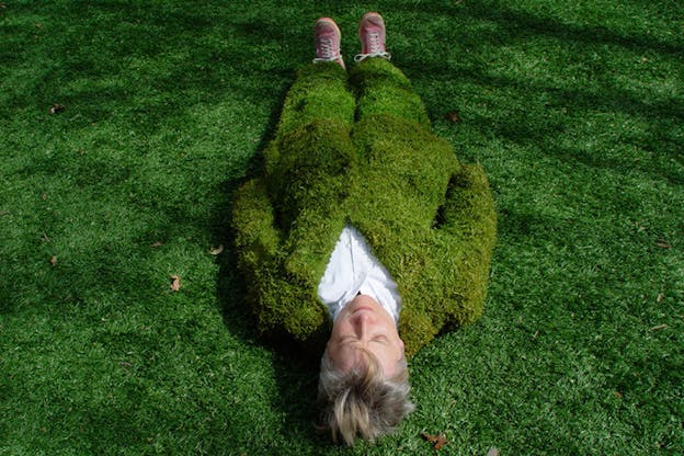 A performer wearing a full body suit made of grass lays on the grass with their eyes closed. A bit of a white shirt peaks through around the neck of the grass suit and the performer wears red sneakers. 