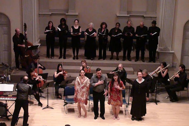 Above angle shot of four performers singing in front of an ensemble and choir. 