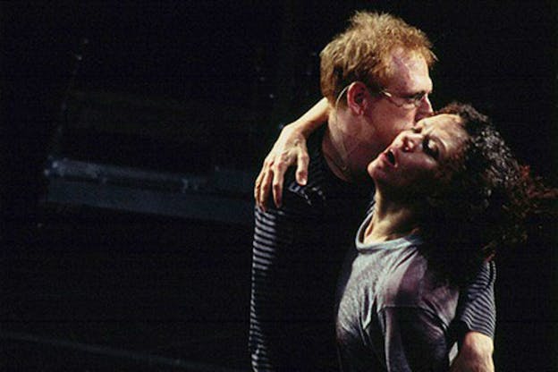 Two people are in front of a dark background. One person, wearing thin wire-rimmed glasses and a striped long sleeve shirt holds Kate Valk who is wearing a grey t-shirt and has short, brown curly hair. Kate Valk appears distressed and has her head tilted back and her mouth slightly open. The person holding her hides their face behind her head. 