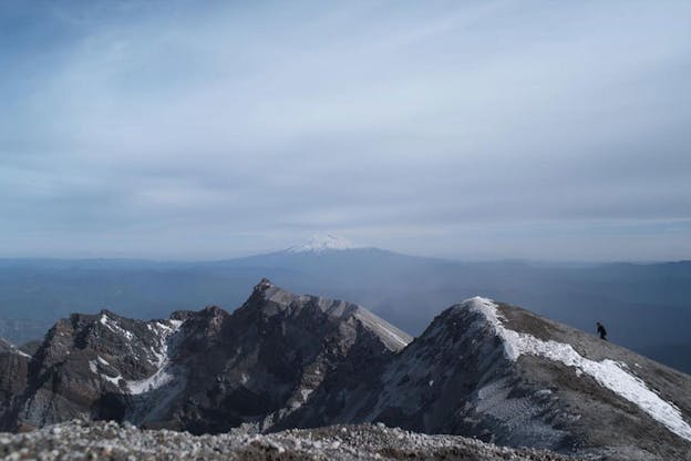 A video still of a snow dusted mountain range. On the right, a small figure is seen climbing these mountains. Behind this, in the distance, the peak of a large mountain is visible.