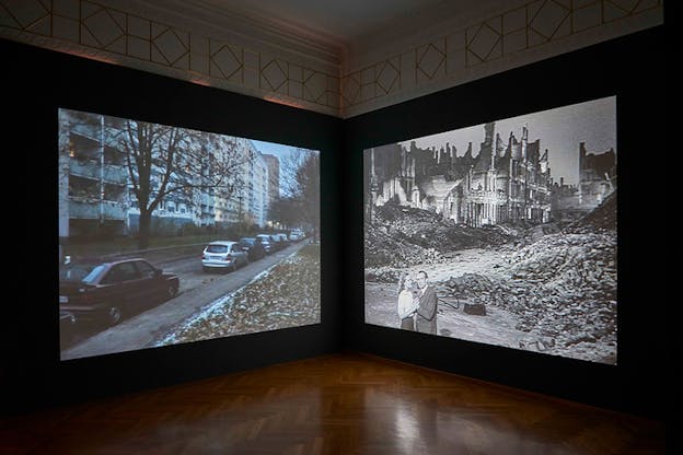Two project installation on two walls. The one on the left depicts apartment buildings with cars in their front streets, the one on the right shows a couple in black and white film holding hands and standing in front of ruins. 