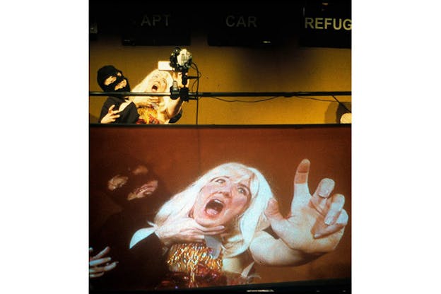 In the upper left, a performer wearing a black ski mask holds the neck of another performer who wears a blonde wig and a gold dress and appears distressed, reaching out towards a camera in front. On the bottom half of this image, this scene between the two performers is projected onto a large screen. 