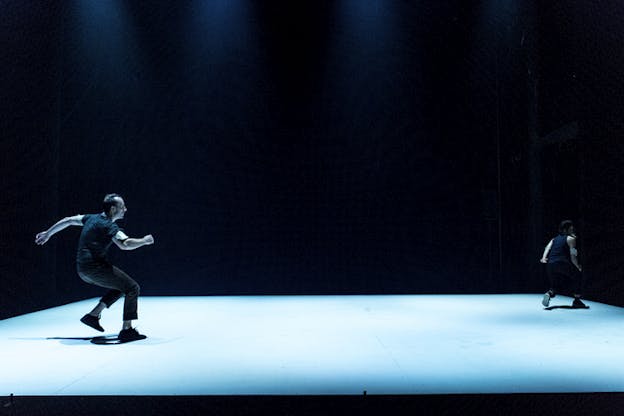 Two performers dressed in black on opposite angles of a square blue lighted floor run towards the same direction. Their surroundings are completely black.