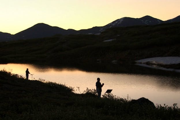 Two black silhouettes with music sheet stands, are situated in front of unmoving water. Behind the water grassy green hills rise with another silhouette turned towards the two people on the other side of the water. A warm yellow sky extends in the back of the hills. 