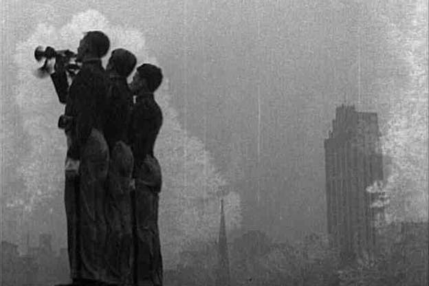 A black and white video still of three people huddled together and looking left, beyond the image's frame. Behind them, there is a large gust of smoke and buildings. 