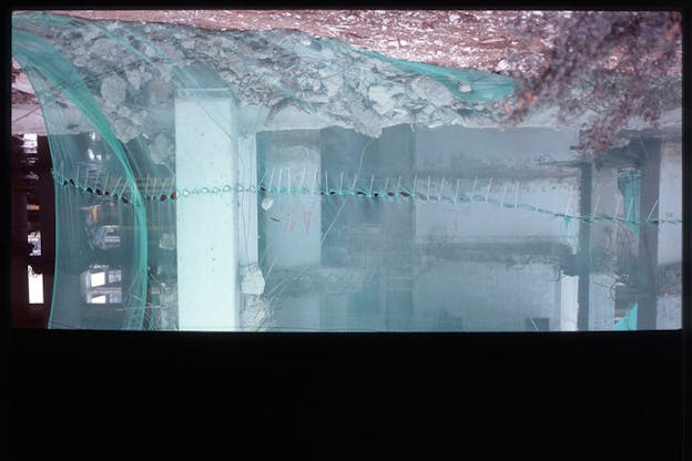 Upside down view of a transparent glacial blue screen divided down the middle with white-strip patched holes covering a pile of rubble in front of a dilapidated building's interiors, and a thick black border along the bottom edge. 