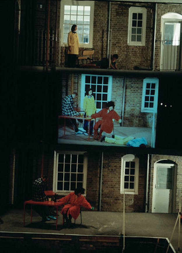 The facade of a staged building contains in its middle a screen projection of four figures, with the one seated and clad in red looking down at a figure who lies face down on the ground. The other two figures watch the scene. Underneath the projection, the same red dressed figure and the one on the ground are siatuated, while on top the two other figures hold the same stances.