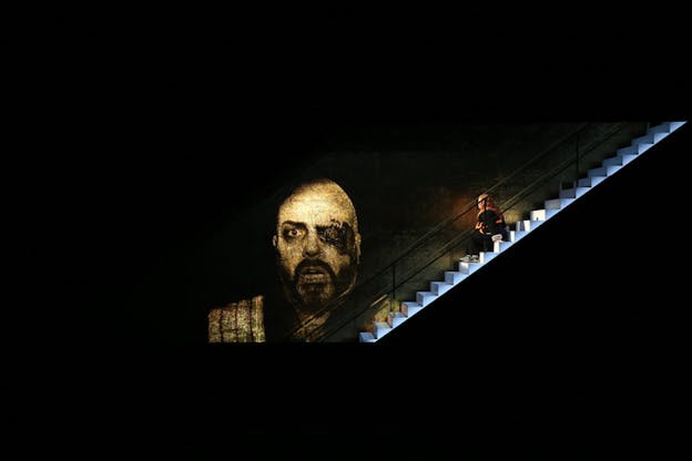 A face frozen in shock is projected on a black stage. Next to it on the right on stairs lighted white sits sideways a figure dressed in black.