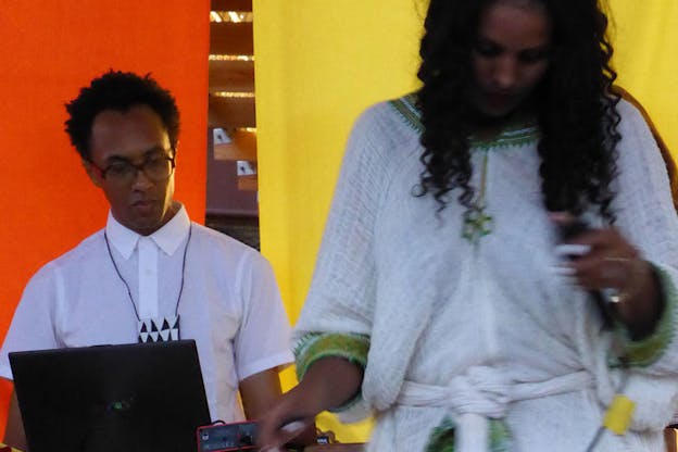 Clayton wearing a white button-up looks at a computer beside a blurred-in-motion person wearing a white and embroidered green blouse, standing in front of two poppy orange and daffodil yellow curtains.