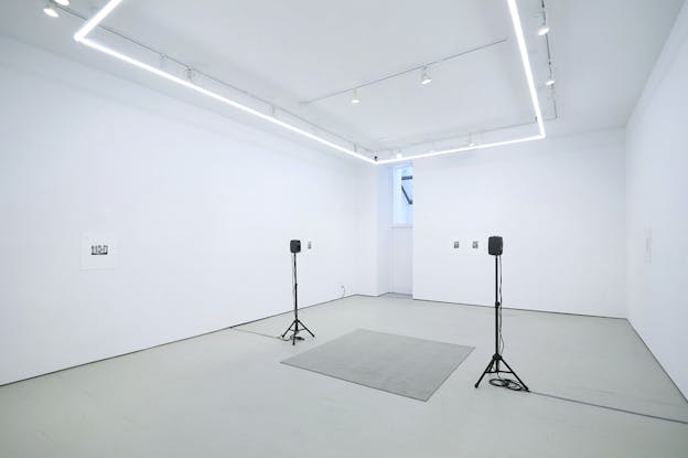 A photo of the inside of a large, brightly lit gallery space taken from a corner of the room. The room is empty except for two small black speakers mounted on tripods that face each other on either side of a square grey rug. The walls are primarily bare with a few small mounted photos.