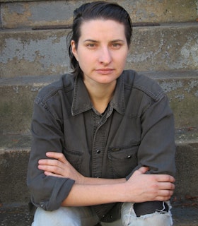 A portrait of MPA sitting in front of a  weathered grey stone wall with her elbows resting on her knees. She has medium length black hair swept to the side and wears a grey collared shirt, and light washed jeans. 