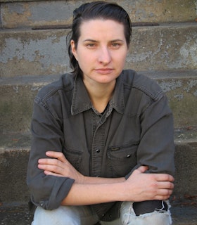 A portrait of MPA sitting in front of a weathered grey stone wall with her elbows resting on her knees. She has medium length black hair swept to the side and wears a grey collared shirt, and light washed jeans.