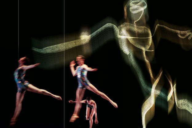 Three performers are in the lower left of a dark srage. They wear leotards with a metallic blue toned abstract pattern on them. Two performers face the left side and leap towards it. A third performer leaps forwards from the background of the stage. A large gold toned sketch of a human figure leaning to the left is projected onto the otherwise black wall behind them. 