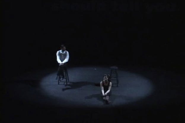 Two performers sit in the center of a spotlight in an otherwise dark, black room. One performer crosses their legs and sits atop a chair and looks at the other performer who sits on the floor, extending their legs outwards, and looking directly forwards. 