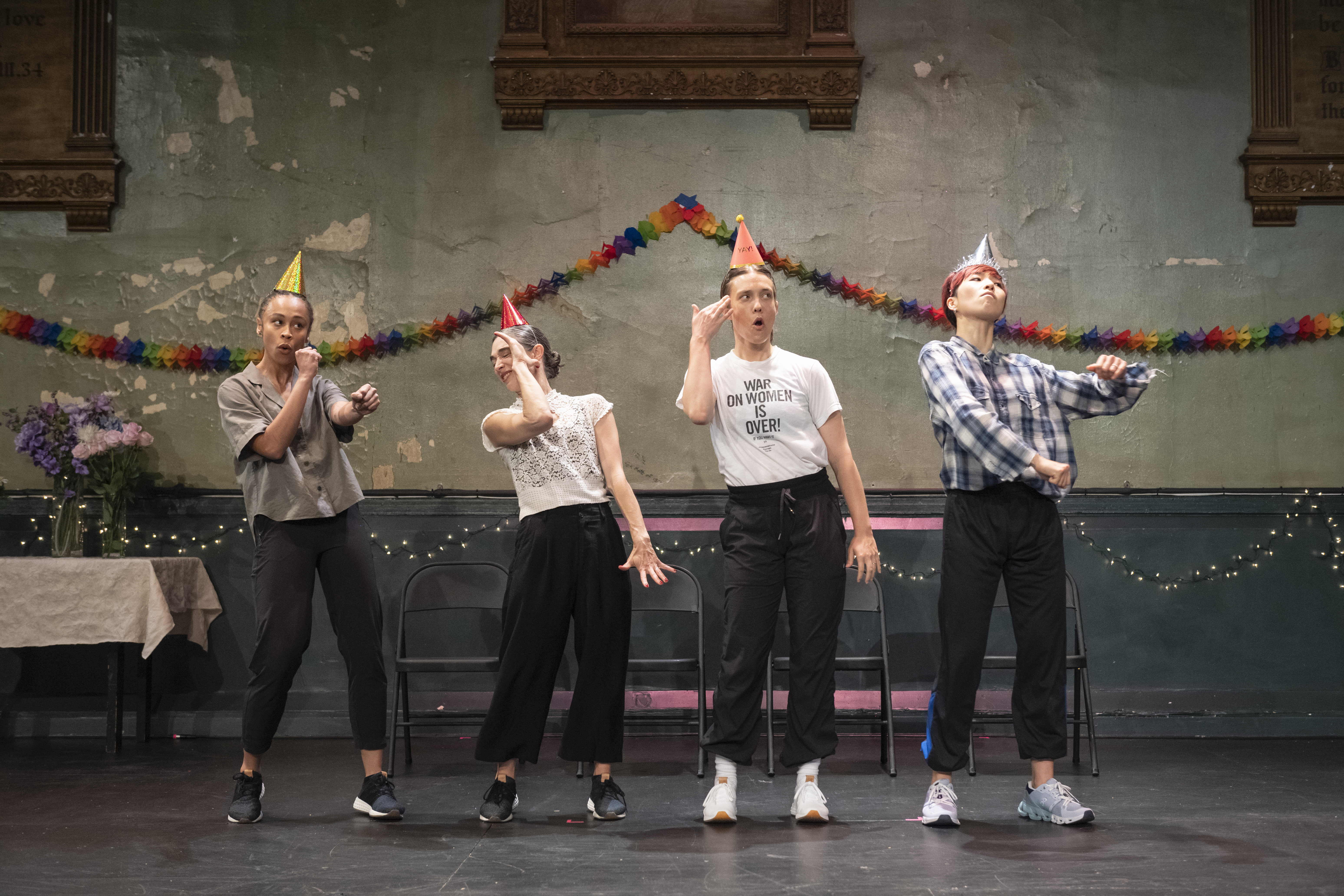 Four performers wearing colorful, cone-shaped party hats stand in a line in various poses in a room decorated for a party. The dancer on the far left, Mykel Marai Nairne, is posed throwing fake punches. Next to her, Monical Bill Barnes is leaning back and averting her face from the camera. Flannery Gregg, on the right half of the frame, stands with her right hand raised to touch her temple, and her left hanging by her side. On the far right, Hsiao-Jou Tang stands with her head tilted back and chin pushed out, gazing out to the right. Her shoulders are leaned back as her arms are curved and rolling in front of her, hands in fists.