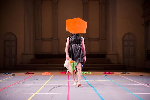 A figure dressed in black leather dress and an orange hexagonal hat covering their face, walks on a catwalk consisting of colorful lines of blue, pink and yellow. 