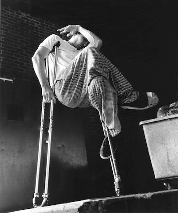 Bill Shannon holds himself off the ground by a crutch under his right arm. His left arm curles over his face while his legs intertwine and hold another crutch.