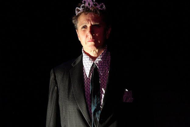 Peggy Shaw wears a black suit jacket, a black tie, a white and black patterned shirt and a plastic tiara. She stands in front of a black background and is lit only from the left side. Pink shadows cover the right side of her body. She has a concerned and preoccupied expression on her face.