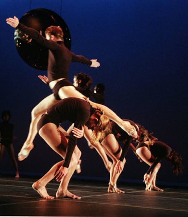 Several performers are situated with a dark blue space. Most of the performers bend forward while one performer leaps over them. All performers wear black long sleeve shirts and black underwear. 