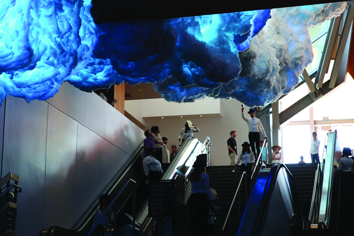 A figure in black pants and a white button accompanied by a black hat, stands on top of stairs with their hand upwards holding a bell. On top of them clouds of white and blue.