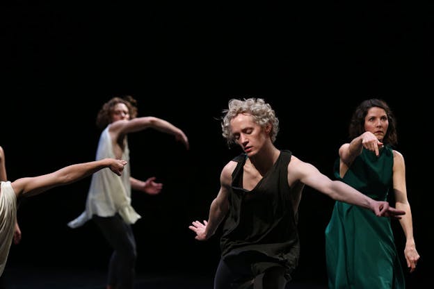 A performer wearing a black tank top stands in the foreground, bending their head down and extending their arms long behind them. In the background, three other performers face the camera and hold out their arms. The performer in the middle holds their arm in a curve at shoulder length in front of their face. The performer on the right holds their arm in front of them and points downwards.