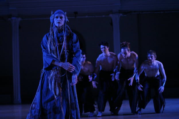 In the foreground, one performer wearing a grey ruffled maxi dress with flare sleeves tilts to the right while gazing to the left side. They wear a hair net headband with floor length braids. In the middle ground, four shirtless perfomers wearing baggy black pants are hunchbacked while arching their arms downward. They all stare at the performer in the front fiercely. In the background, two columns are placed against a black wall. The stage is tinged with cool toned lights.