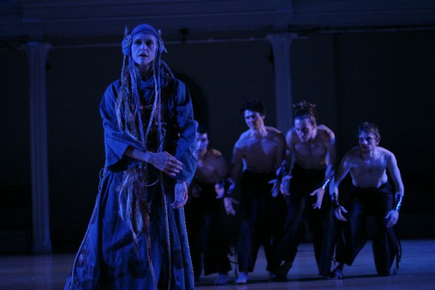 In the foreground, one performer wearing a grey ruffled maxi dress with flare sleeves tilts to the right while gazing to the left side. They wear a hair net headband with floor length braids. In the middle ground, four shirtless perfomers wearing baggy black pants are hunchbacked while arching their arms downward. They all stare at the performer in the front fiercely. In the background, two columns are placed against a black wall. The stage is tinged with cool toned lights.