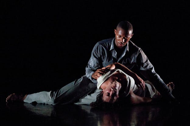 A man seated on the ground looks down touching the woman lying down next to him with her eyes closed. Both performers are surrounded by a black background.