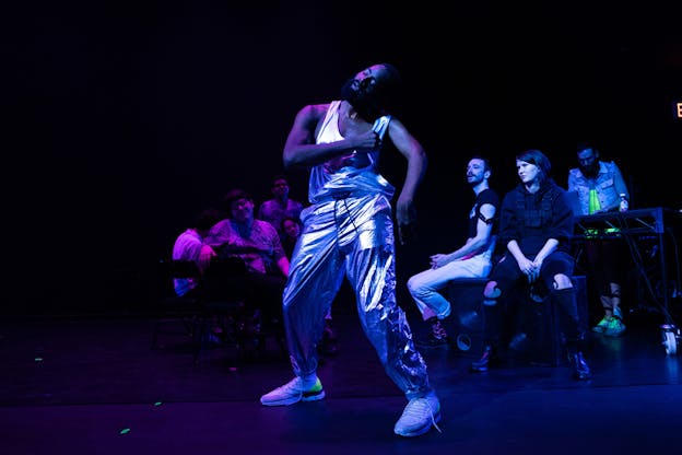 A performer dressed in silver metallic pants dances with his eyes closed as  people sit behind him on the stage. They are all illuminated by a pink and purple light.