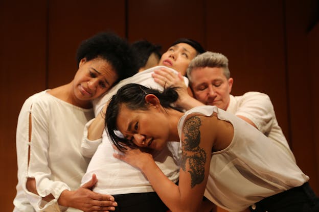 Five performers embrace in a huddle, their faces displaying a range of emotions from care to comfort to concern. They are all wearing white shirts.